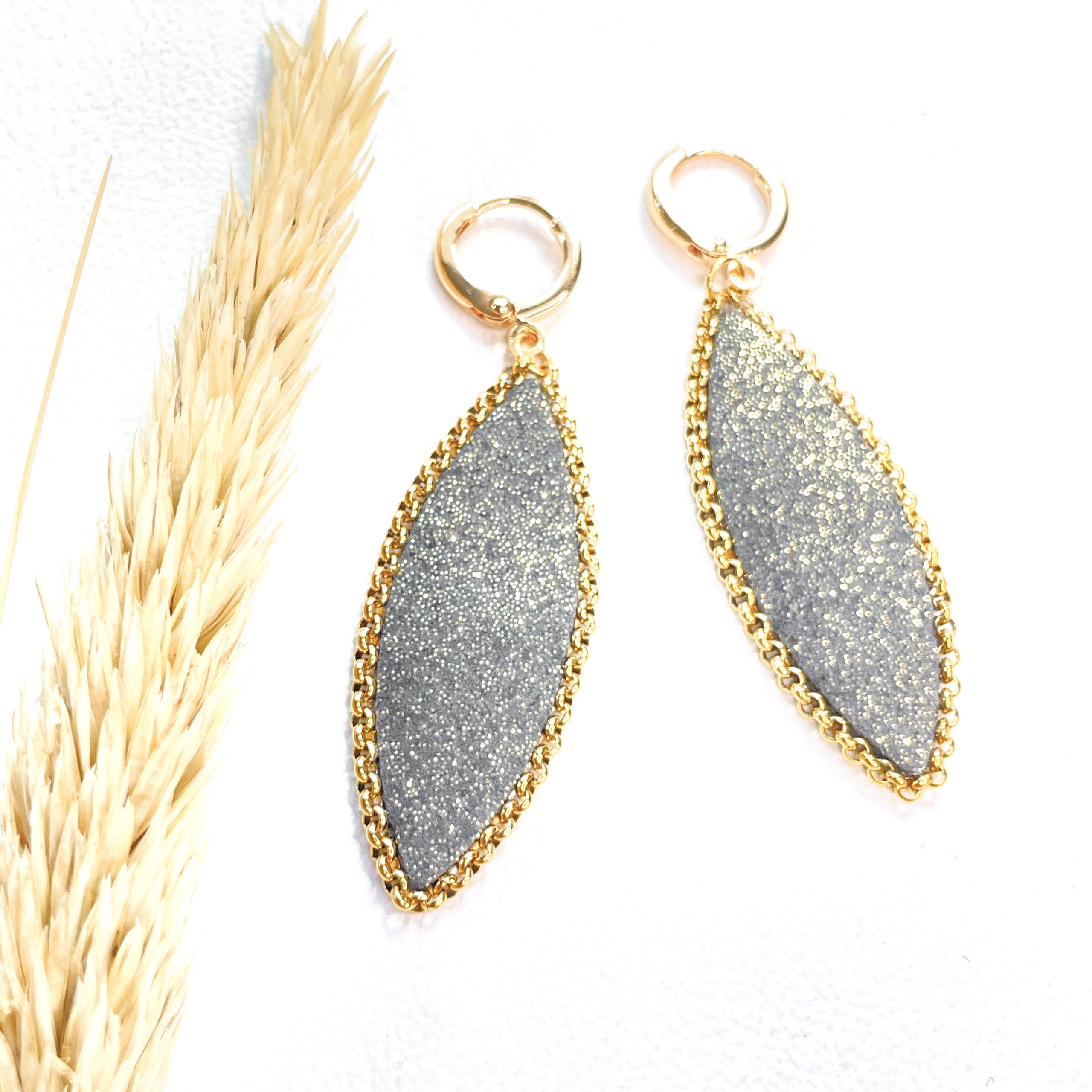 Paire de boucles d'oreilles pendantes Nune modèle Auris en forme d'épi en cuir gris pailleté or cerclé de chaîne dorée, sur fond blanc avec épi