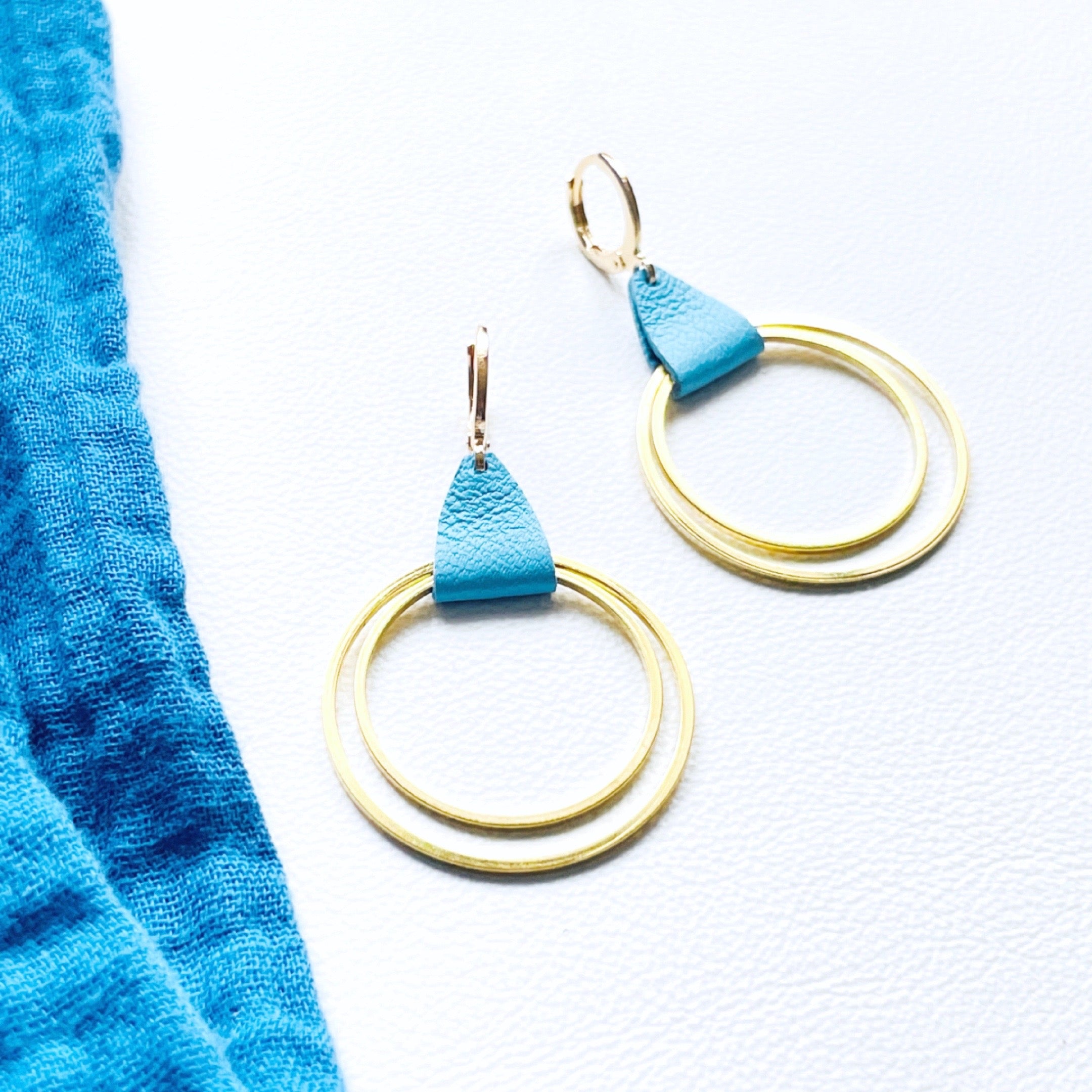 Créoles dorées du modèle Vega de la marque Nune, avec une touche de cuir bleu pastel, présentées sur fond blanc avec un tissu de la même teinte