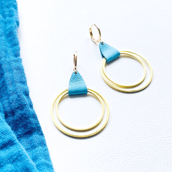 Créoles dorées du modèle Vega de la marque Nune, avec une touche de cuir bleu pastel, présentées sur fond blanc avec un tissu de la même teinte