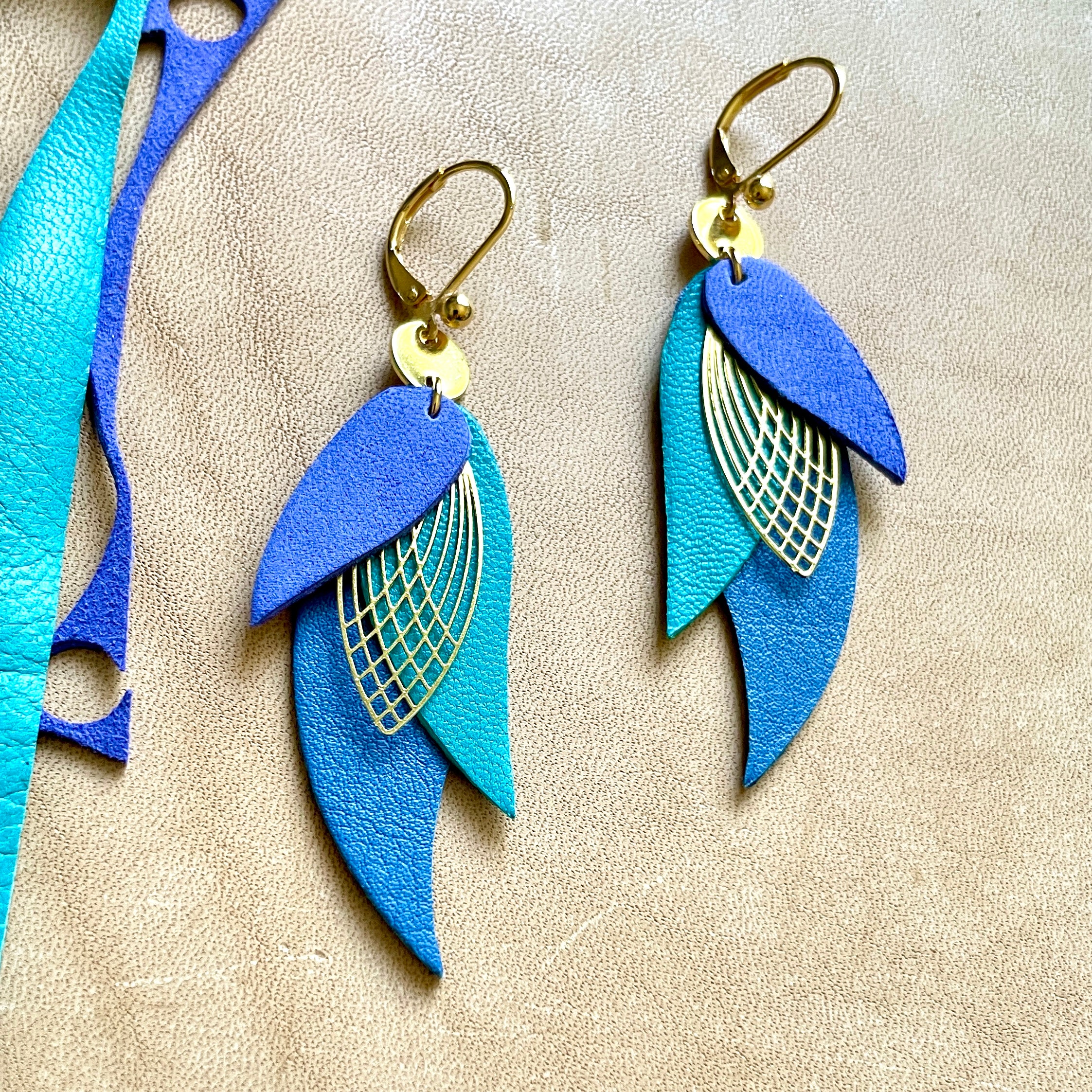 boucles d'oreilles dorées Nune modèle Ara en plumes de cuir violet velours, bleu turquoise et bleu denim mat sur cuir beige