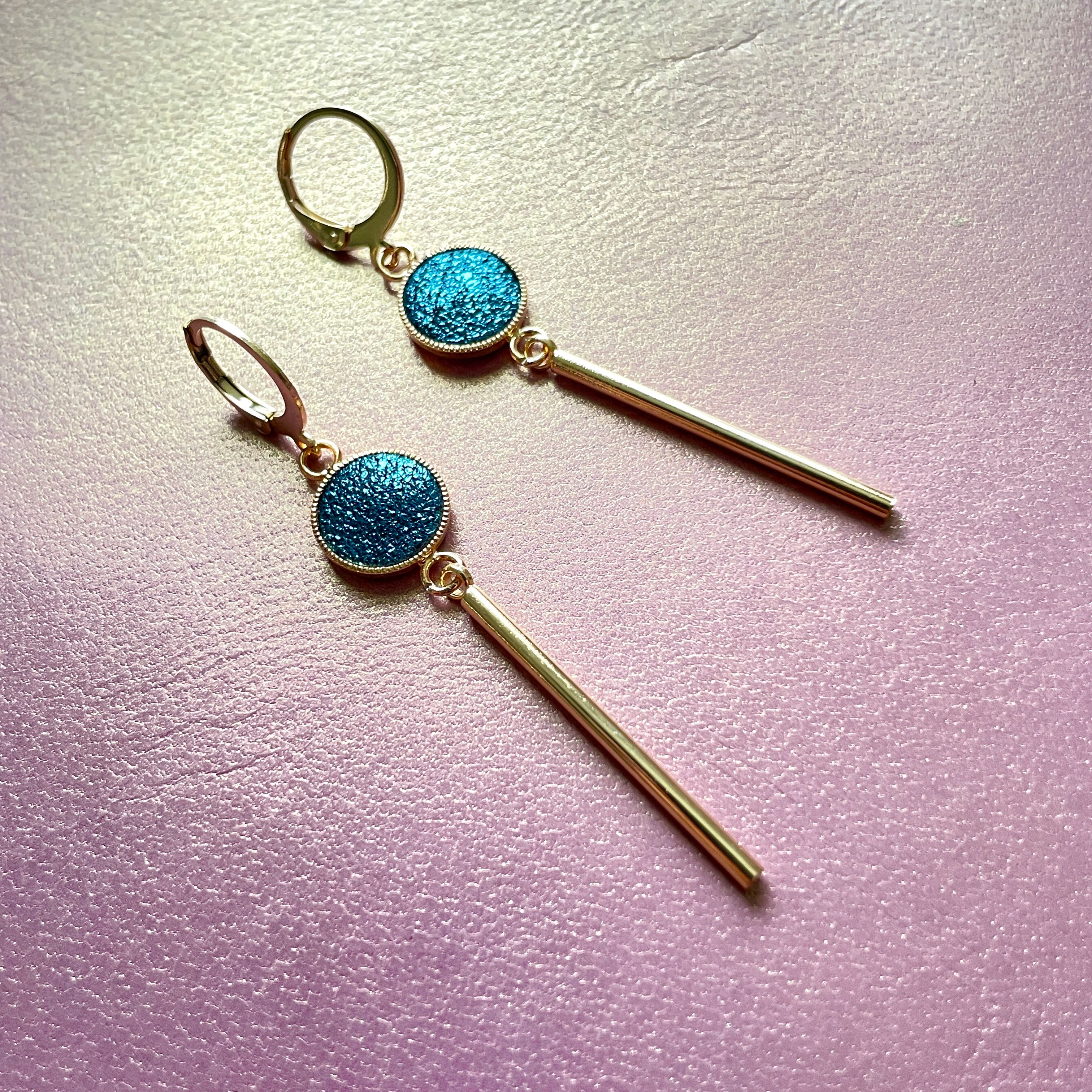 Nune longues et fines boucles d'oreilles dorées Nida avec serti en cuir bleu paon pailleté, sur fond cuir mauve irisé