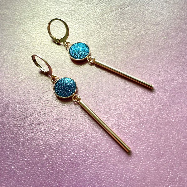 Nune longues et fines boucles d'oreilles dorées Nida avec serti en cuir bleu paon pailleté, sur fond cuir mauve irisé