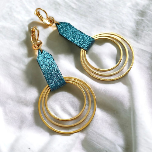 Grandes boucles d'oreilles dorées avec pendentifs en anneaux concentriques attachés par des rubans de cuir recyclé bleu paon pailleté, sur un tissu blanc. Modèle Dizzo