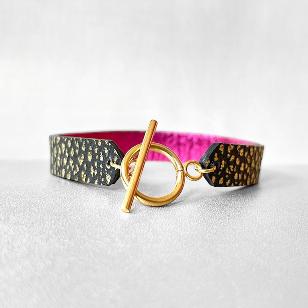 Bracelet en cuir double face bicolore, avec cuir fushia métallisé au verso et cuir noir grainé or au recto avec fermoir boucle dorée en T, sur fond gris
