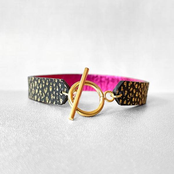 Bracelet en cuir double face bicolore, avec cuir fushia métallisé au verso et cuir noir grainé or au recto avec fermoir boucle dorée en T, sur fond gris