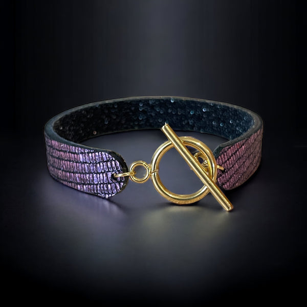 Bracelet en cuir double face bicolore, avec cuir violet brillant façon serpent au recto et cuir noir grainé pailleté au verso avec fermoir boucle dorée en T, sur fond noir
