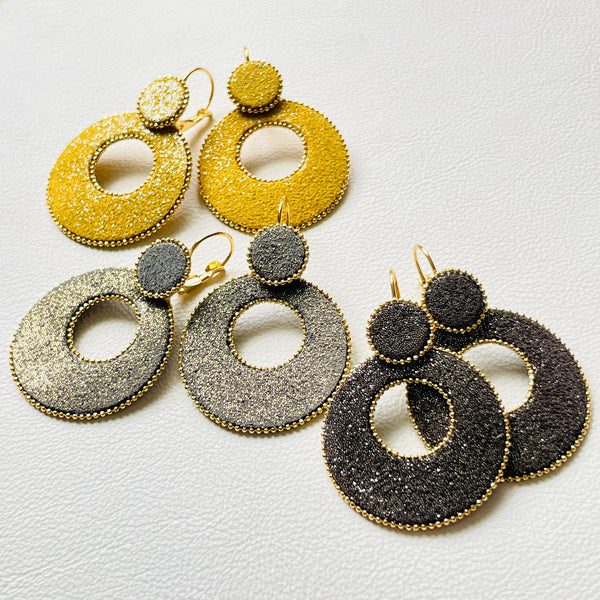 boucles d'oreilles créoles Nune modèle Céleste en forme de cercles en cuir gris pailleté or, safran pailleté argent et anthracite pailleté argent cerclées de chaîne dorée, sur fond cuir blanc