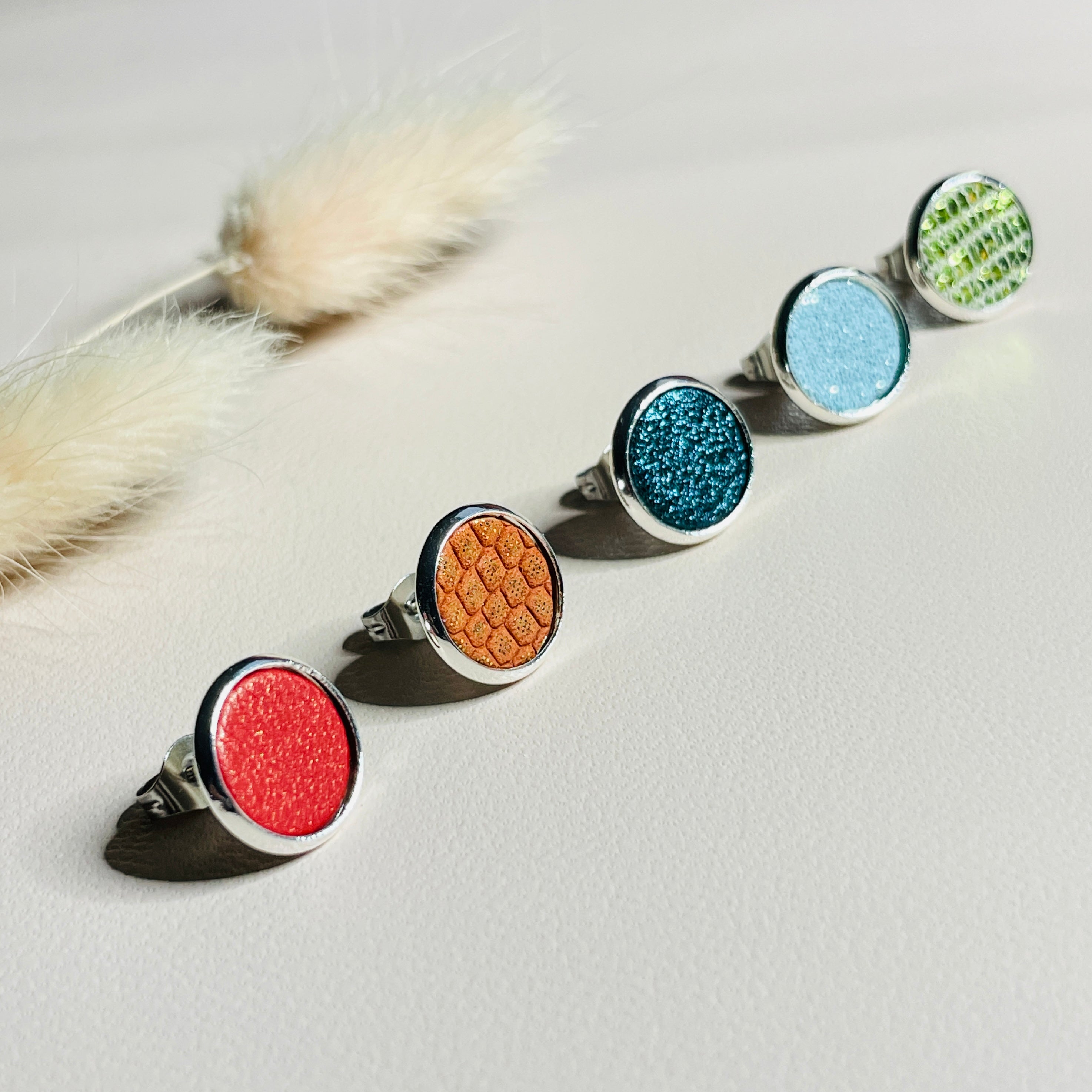 Puces d'oreilles argentées rondes Nune modèle Ilis argenté dans 5 coloris de cuirs coquelicot nacré, orange or façon serpent, saphir pailleté, topaze pailleté et vert olive métallisé, sur fond de cuir blanc cassé