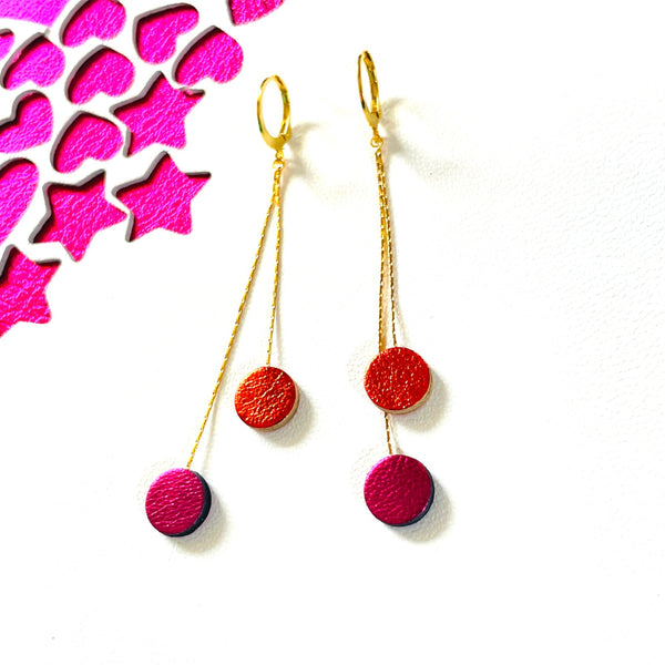 Longues et fines boucles d'oreilles Nune dorées terminées petits disques de cuir recyclé orange sanguine métallisé et fushia métallisé, sur fond blanc et fushia étoilé. Modèle Merry