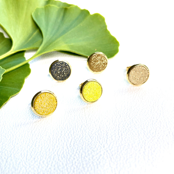 Puces d'oreilles dorées rondes Nune modèle Ilis dans 5 coloris de cuirs pailletés dans les tons jaunes, sur fond blanc avec feuilles de Gingko