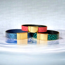 Charger l&#39;image dans la galerie, 3 bracelets en cuir marin rose saumon, bleu saphir métallisé et vert émeraude écaille avec fermoirs dorés aimantés, posés sur l&#39;eau
